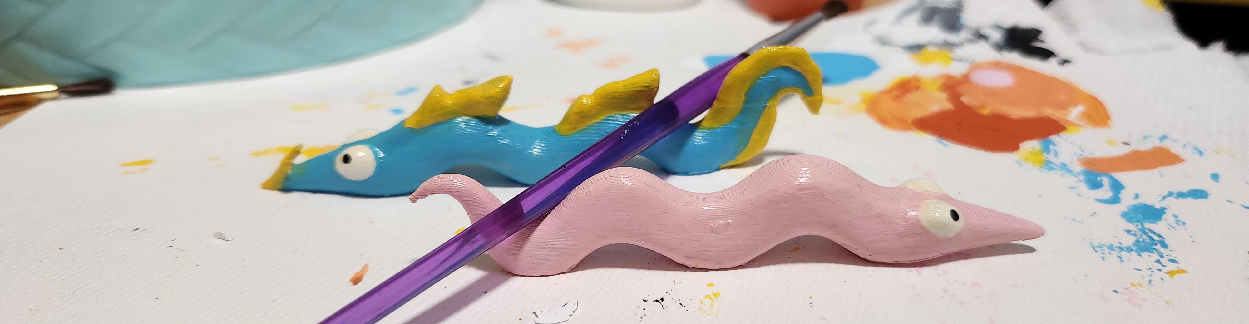 two brush rests, one painted like a pink worm on string, the other a ribbon eel